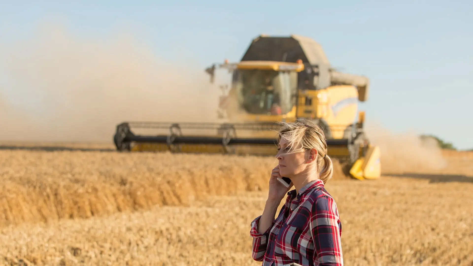 Female farmer on mobile phone in wheat field with harvester in background – agricultural connectivity and rural communications