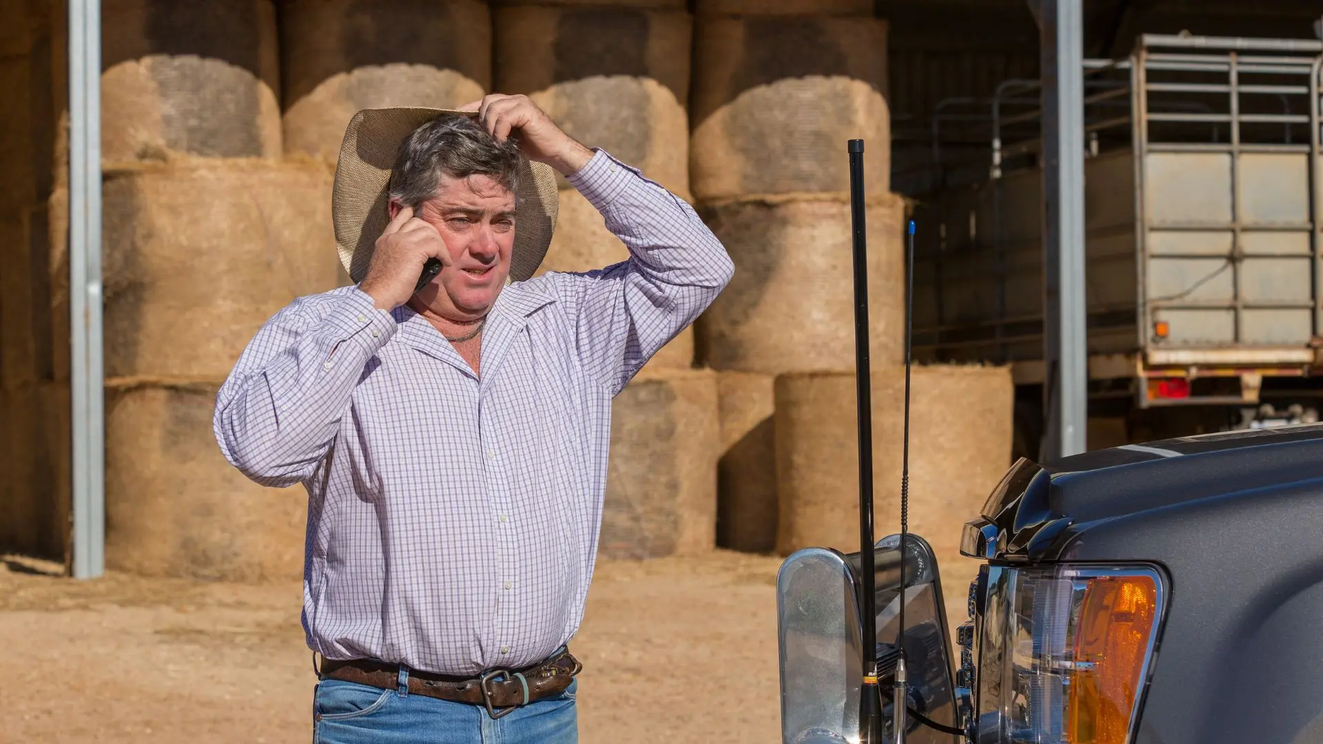 Farmer using mobile phone near hay bales with 4WD vehicle antenna, highlighting rural communication challenges