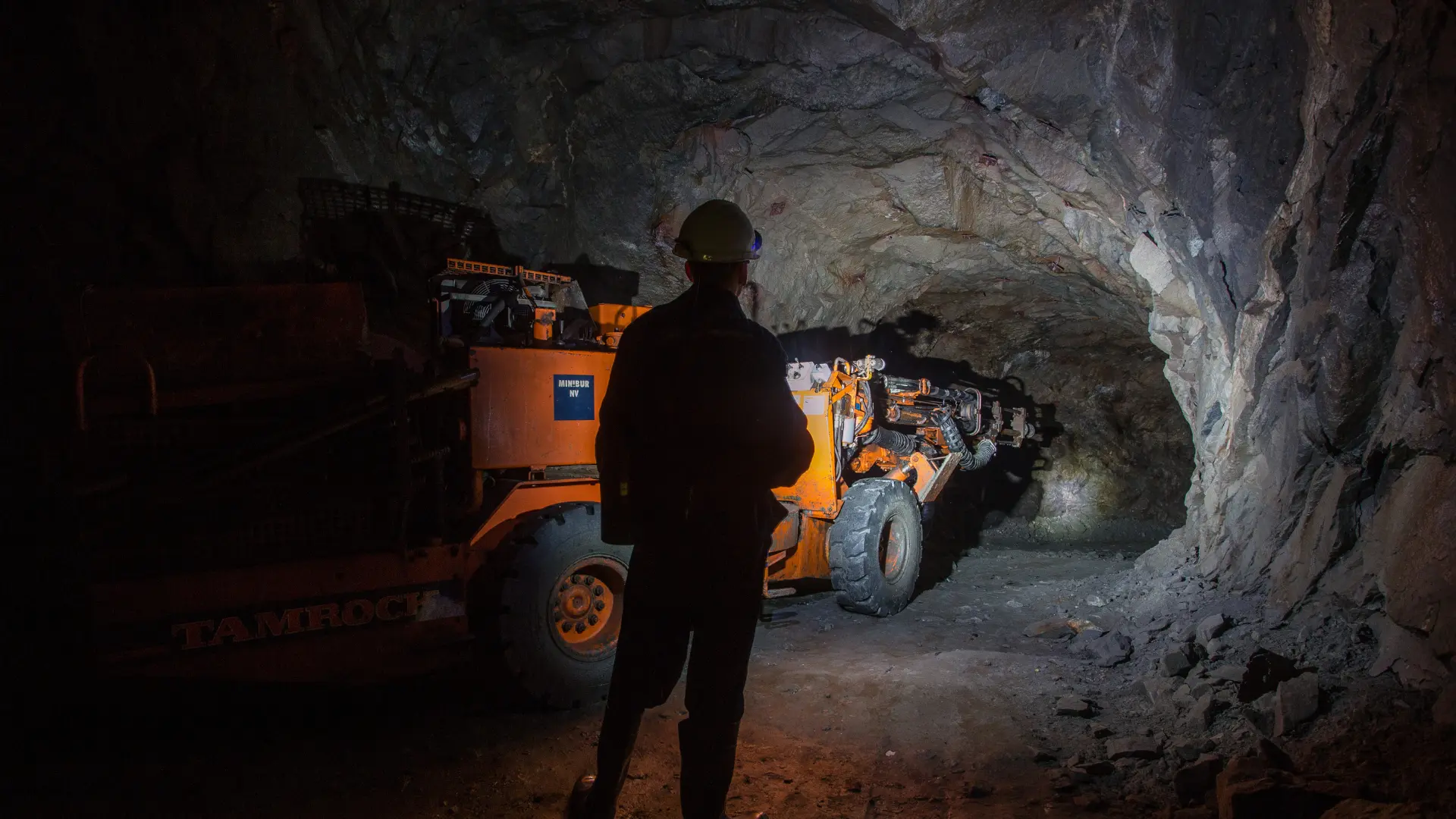 A miner wearing a helmet and reflective clothing stands in an underground tunnel illuminated by artificial light. An orange mining vehicle operates deeper inside the dark, rocky tunnel.