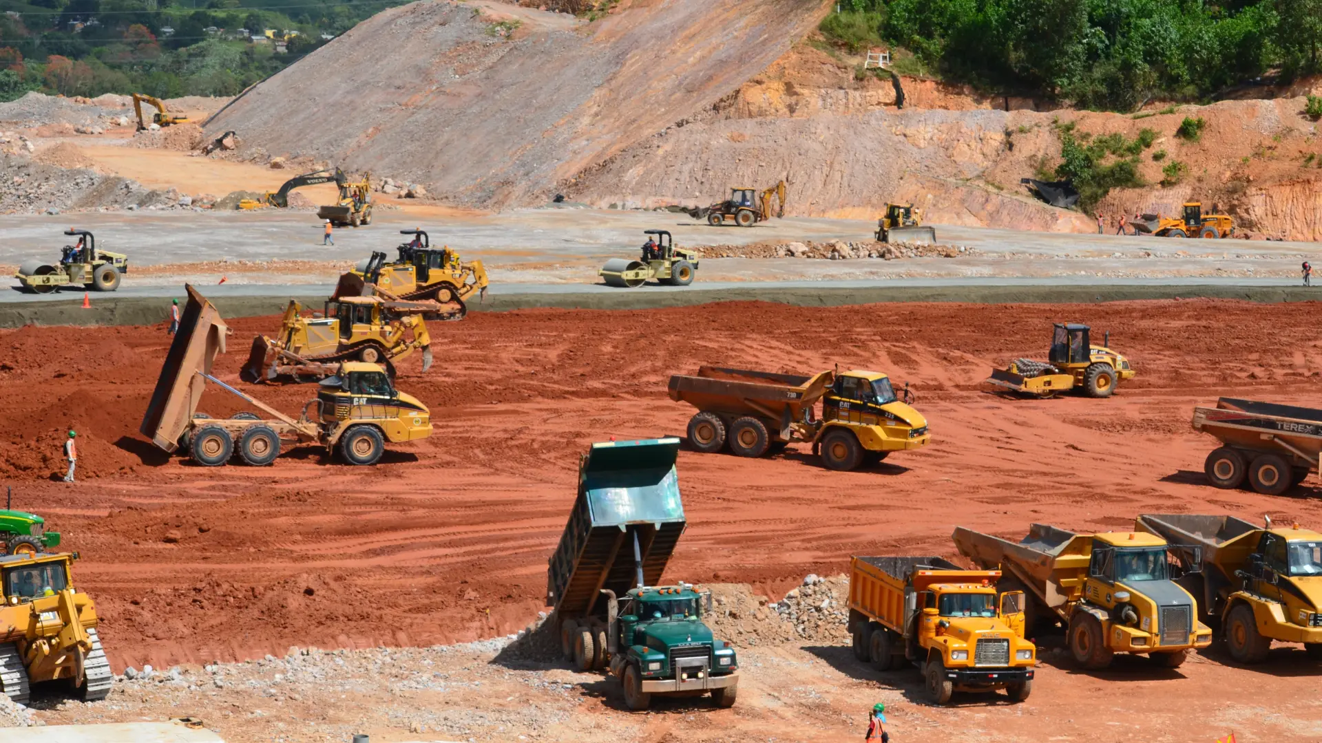 Large-scale open-pit mining site with multiple dump trucks, excavators, and bulldozers working on red soil. Machinery is actively loading and transporting earth, with operators visible across the site.