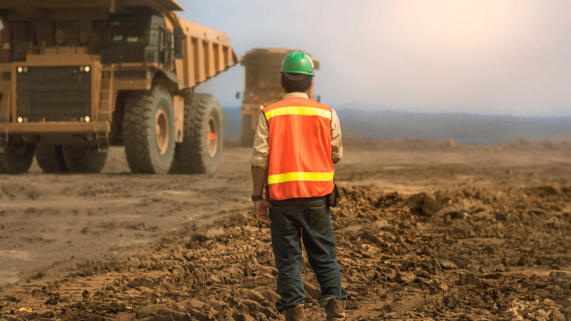 Construction or mining worker wearing a safety vest and helmet stands on a muddy site, observing massive haul trucks operating in the distance under hazy conditions.